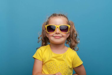 Kıvırcık saçlı, sarı gözlüklü ve sarı tişörtlü neşeli bir kız çocuğu, neşeli bir stüdyo resminde parlak mavi bir arka plana karşı güvenle poz veriyor..
