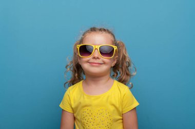 Kıvırcık saçlı, sarı gözlüklü ve sarı tişörtlü neşeli bir kız çocuğu, neşeli bir stüdyo resminde parlak mavi bir arka plana karşı güvenle poz veriyor..