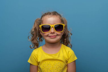 Kıvırcık saçlı, sarı gözlüklü ve sarı tişörtlü neşeli bir kız çocuğu, neşeli bir stüdyo resminde parlak mavi bir arka plana karşı güvenle poz veriyor..