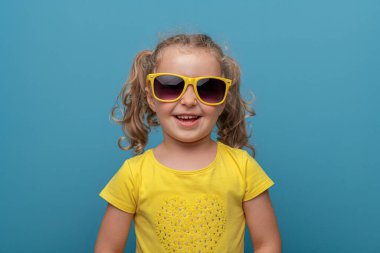 Kıvırcık saçlı, sarı gözlüklü ve sarı tişörtlü neşeli bir kız çocuğu, neşeli bir stüdyo resminde parlak mavi bir arka plana karşı güvenle poz veriyor..