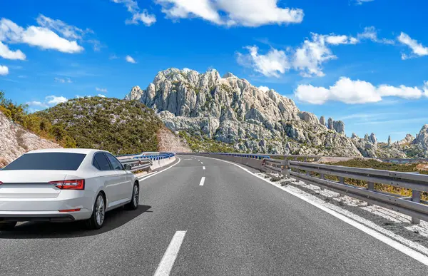 Beyaz bir sedan, sarmal asfalt bir yolda bariyerlerle ilerliyor. Parlak mavi bulutların altında engebeli bir dağ zirvesine doğru ilerliyor..