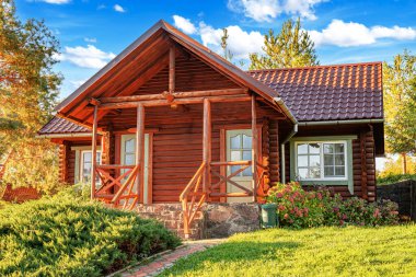 Kırsal ahşap bir kulübe kiremit çatılı ve taş temeli yeşil, çiçek ve ağaçlarla çevrili parlak bir yaz gökyüzü altında.