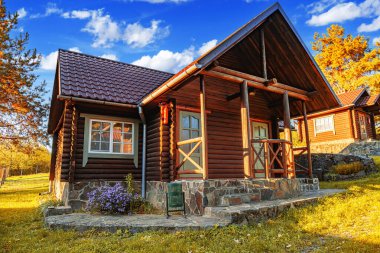 Kırsal ahşap bir kulübe kiremit çatılı ve taş temeli yeşil, çiçek ve ağaçlarla çevrili parlak bir yaz gökyüzü altında.