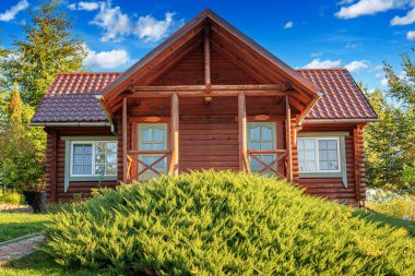 Kırsal ahşap bir kulübe kiremit çatılı ve taş temeli yeşil, çiçek ve ağaçlarla çevrili parlak bir yaz gökyüzü altında.