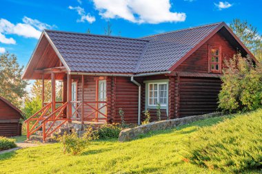 Kırsal ahşap bir kulübe kiremit çatılı ve taş temeli yeşil, çiçek ve ağaçlarla çevrili parlak bir yaz gökyüzü altında.