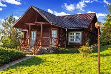 Kırsal ahşap bir kulübe kiremit çatılı ve taş temeli yeşil, çiçek ve ağaçlarla çevrili parlak bir yaz gökyüzü altında.