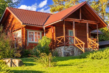 Kırsal ahşap bir kulübe kiremit çatılı ve taş temeli yeşil, çiçek ve ağaçlarla çevrili parlak bir yaz gökyüzü altında.