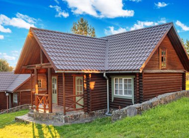 Kırsal ahşap bir kulübe kiremit çatılı ve taş temeli yeşil, çiçek ve ağaçlarla çevrili parlak bir yaz gökyüzü altında.