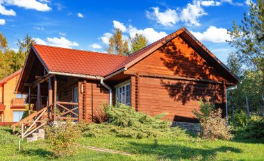 Kırsal ahşap bir kulübe kiremit çatılı ve taş temeli yeşil, çiçek ve ağaçlarla çevrili parlak bir yaz gökyüzü altında.
