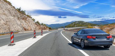 Gri bir coupe, parlak mavi gökyüzünün altında dolambaçlı bir dağ yolu boyunca gidiyor. Dağılmış bulutlar, yaz yolculuğunun özünü yakalıyor..