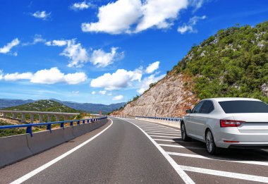 Beyaz bir sedan dolambaçlı bir dağ yolunun kenarına park edilmiş. Parlak mavi bir gökyüzünün altında. Dağılmış bulutlar, güzel bir yaz yolculuğu sahnesi sergiliyor..