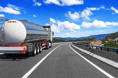 Bir tanker modern bir otoyol boyunca mavi gökyüzü ve bulutlarla dolu manzaralı bir kırsal bölgeden geçiyor..