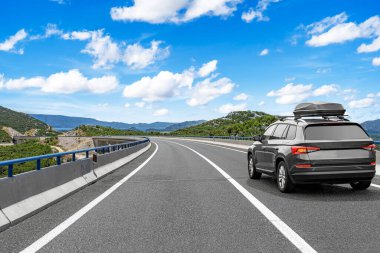 Çatı kutusu olan modern bir SUV, bulutlu mavi bir gökyüzünün altında manzaralı bir dağ manzarası boyunca yol alır..