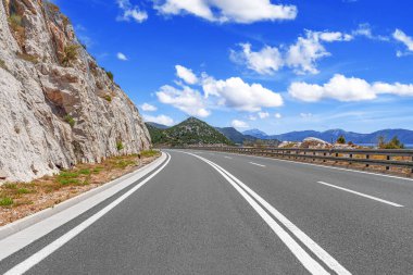 Boş, dolambaçlı bir yol sarp, kayalık bir dağın etrafında kıvrılır parlak mavi bir gökyüzünün altında dağınık bulutlarla..