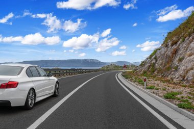 Beyaz bir araba, parlak mavi bir gökyüzünün altında, tüylü bulutlarla manzaralı bir dağ yolunda ilerliyor..