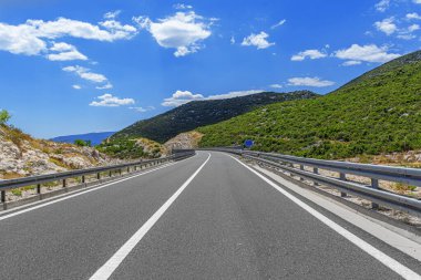 Boş, dolambaçlı bir yol, beyaz bulutlu açık mavi gökyüzünün altında dağlık bir arazide kıvrılır..