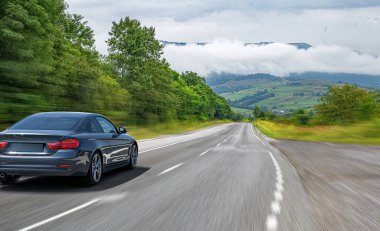 Pürüzsüz, siyah bir coupe araba yeşil tarlaları, ağaçları ve arka planda sisli dağları olan manzaralı kırsal bir yolda hızla ilerliyor..