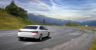 Beyaz bir sedan, yeşil tepeler ve bulutlu mavi gökyüzü ile çevrili dolambaçlı bir dağ yolunda ilerliyor..