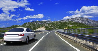 A white car drives along a mountain highway on a bright sunny day with blue sky and clouds, symbolizing travel, freedom, and adventure.