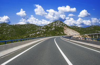 Modern bir otoyol, yemyeşil, kayalık tepeler ve kabarık bulutlarla dolu parlak mavi gökyüzü ile dağlık bir arazide kıvrılır..