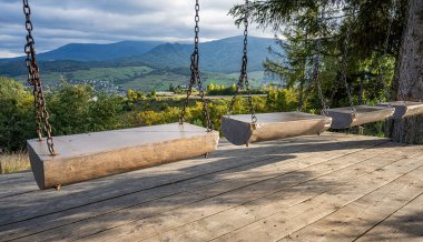 Platformda kırsal bir ahşap salıncak, güneşli bir günde dağları, ormanları ve uzak bir köyü gören manzaralı bir vadiye bakıyor..