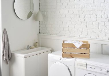 Beyaz banyo, çamaşır ve kuru makine, tuğla duvar ve şeffaf ekran, dolap lavabo ve ayna, bitki, kirli çamaşırlar.