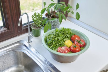 Bir kadın mutfak lavabosunda taze limon, domates ve yapraklı sebzeleri yıkıyor. Güneşli bir pencerenin yanında sağlıklı malzemeler hazırlıyor..