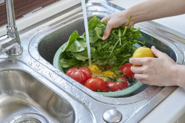 Bir kadın mutfak lavabosunda taze limon, domates ve yapraklı sebzeleri yıkıyor. Güneşli bir pencerenin yanında sağlıklı malzemeler hazırlıyor..
