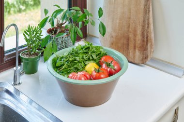 Bir kadın mutfak lavabosunda taze limon, domates ve yapraklı sebzeleri yıkıyor. Güneşli bir pencerenin yanında sağlıklı malzemeler hazırlıyor..