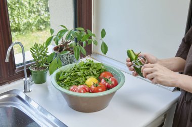Bir kadın mutfak lavabosunda taze limon, domates ve yapraklı sebzeleri yıkıyor. Güneşli bir pencerenin yanında sağlıklı malzemeler hazırlıyor..