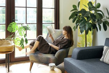 Bir kadın, geniş bir pencerenin yanında, bir koltukta rahatlar, bir kitap okumaya dalmış, yemyeşil bitkilerle çevrili..