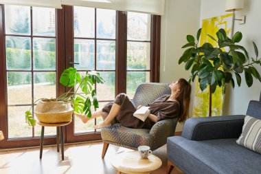 Bir kadın, geniş bir pencerenin yanında, bir koltukta rahatlar, bir kitap okumaya dalmış, yemyeşil bitkilerle çevrili..