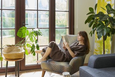 Bir kadın, geniş bir pencerenin yanında, bir koltukta rahatlar, bir kitap okumaya dalmış, yemyeşil bitkilerle çevrili..