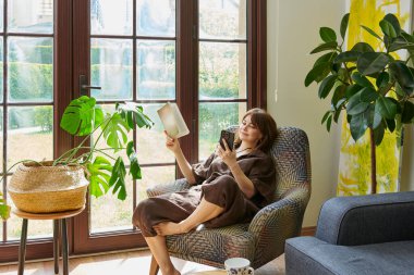 Bir kadın, geniş bir pencerenin yanında, bir koltukta rahatlar, bir kitap okumaya dalmış, yemyeşil bitkilerle çevrili..