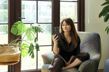 Bir kadın, geniş bir pencerenin yanında, bir koltukta rahatlar, bir kitap okumaya dalmış, yemyeşil bitkilerle çevrili..