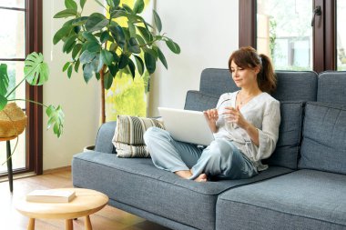 Bir kadın evinde rahat bir şekilde kanepede çalışır, bilgisayarını kullanır ve telefonda konuşur, bitkilerle ve doğal ışıkla çevrili..
