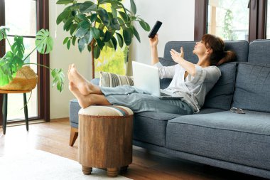 Bir kadın evinde rahat bir şekilde kanepede çalışır, bilgisayarını kullanır ve telefonda konuşur, bitkilerle ve doğal ışıkla çevrili..