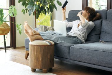 Bir kadın evinde rahat bir şekilde kanepede çalışır, bilgisayarını kullanır ve telefonda konuşur, bitkilerle ve doğal ışıkla çevrili..