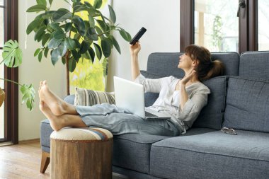 Bir kadın evinde rahat bir şekilde kanepede çalışır, bilgisayarını kullanır ve telefonda konuşur, bitkilerle ve doğal ışıkla çevrili..