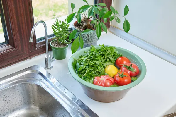 Bir kadın mutfak lavabosunda taze limon, domates ve yapraklı sebzeleri yıkıyor. Güneşli bir pencerenin yanında sağlıklı malzemeler hazırlıyor..