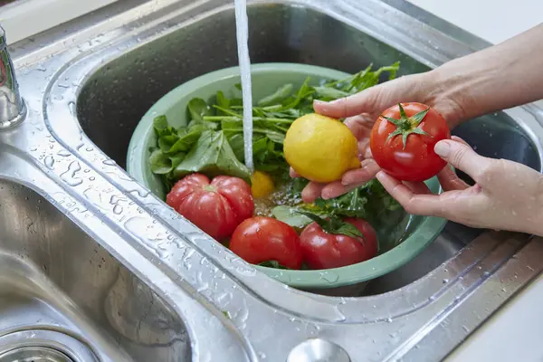 Bir kadın mutfak lavabosunda taze limon, domates ve yapraklı sebzeleri yıkıyor. Güneşli bir pencerenin yanında sağlıklı malzemeler hazırlıyor..