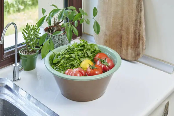 Bir kadın mutfak lavabosunda taze limon, domates ve yapraklı sebzeleri yıkıyor. Güneşli bir pencerenin yanında sağlıklı malzemeler hazırlıyor..