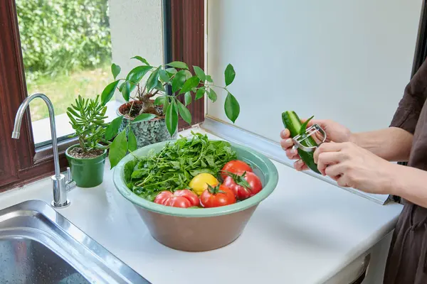 Bir kadın mutfak lavabosunda taze limon, domates ve yapraklı sebzeleri yıkıyor. Güneşli bir pencerenin yanında sağlıklı malzemeler hazırlıyor..