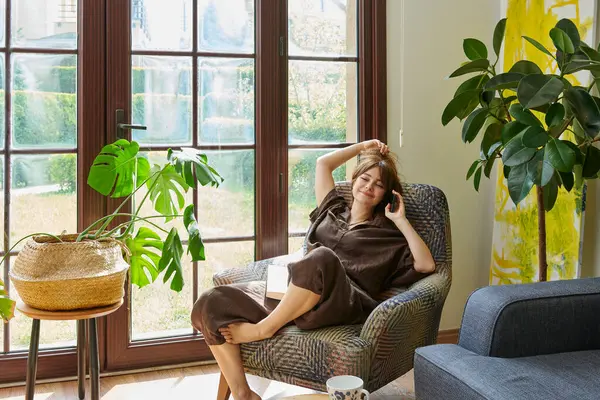 Bir kadın, geniş bir pencerenin yanında, bir koltukta rahatlar, bir kitap okumaya dalmış, yemyeşil bitkilerle çevrili..