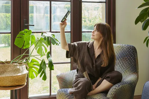 Bir kadın, geniş bir pencerenin yanında, bir koltukta rahatlar, bir kitap okumaya dalmış, yemyeşil bitkilerle çevrili..