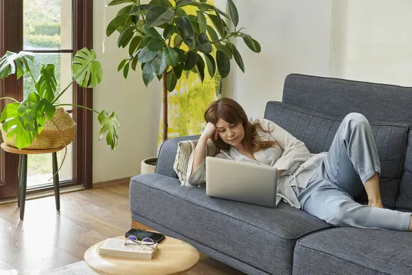 Bir kadın evinde rahat bir şekilde kanepede çalışır, bilgisayarını kullanır ve telefonda konuşur, bitkilerle ve doğal ışıkla çevrili..