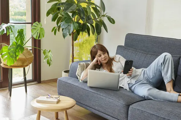 Bir kadın evinde rahat bir şekilde kanepede çalışır, bilgisayarını kullanır ve telefonda konuşur, bitkilerle ve doğal ışıkla çevrili..