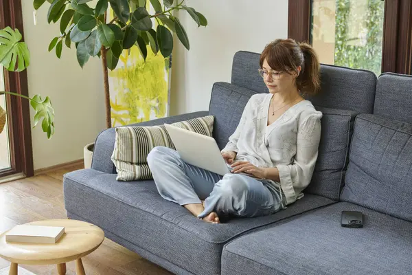 Bir kadın evinde rahat bir şekilde kanepede çalışır, bilgisayarını kullanır ve telefonda konuşur, bitkilerle ve doğal ışıkla çevrili..