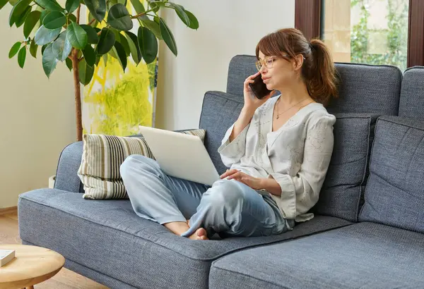 Bir kadın evinde rahat bir şekilde kanepede çalışır, bilgisayarını kullanır ve telefonda konuşur, bitkilerle ve doğal ışıkla çevrili..