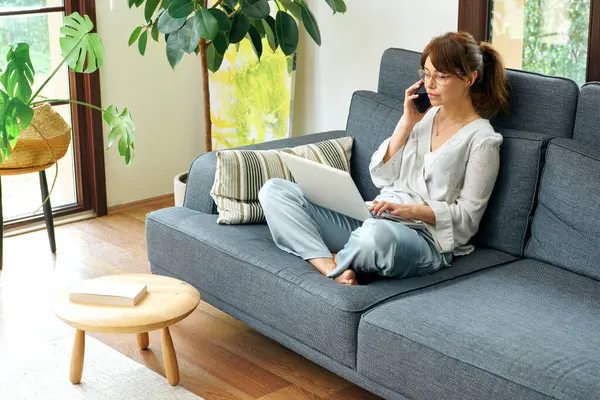 Bir kadın evinde rahat bir şekilde kanepede çalışır, bilgisayarını kullanır ve telefonda konuşur, bitkilerle ve doğal ışıkla çevrili..